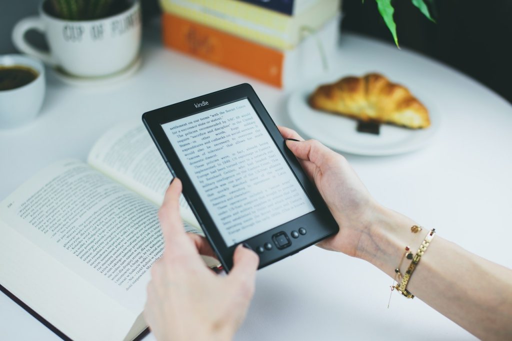 mão aparentemente femininas segurando um amazon kindle, ao fundo é possível ver uma mesa com um livro em cima