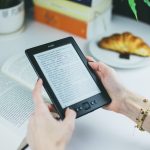 mão aparentemente femininas segurando um amazon kindle, ao fundo é possível ver uma mesa com um livro em cima