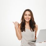 amazing businesswoman holding silver laptop pointing finger sideways with smile white wall min