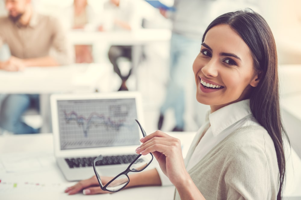 mulher sorrindo com óculos na mão na frente de um notebook para ilustrar texto sobre período de experiência no trabalho